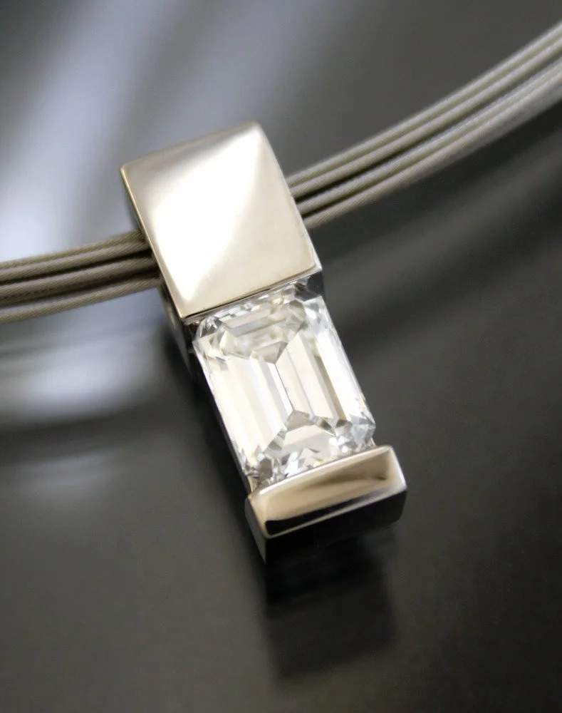 A modern silver necklace with a rectangular, clear gemstone pendant resting on a black surface.