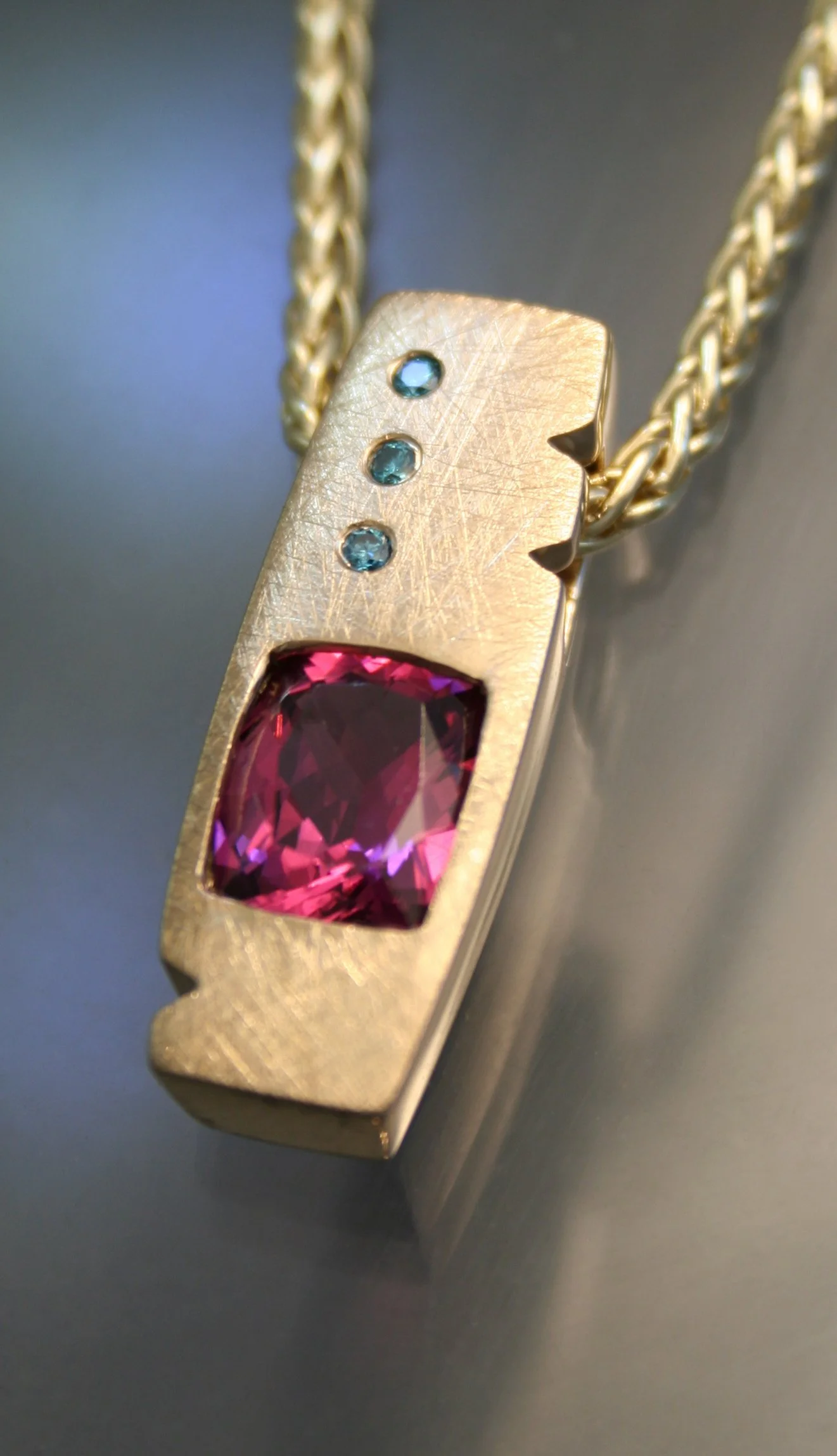 Gold necklace pendant with a large pink gemstone and small blue gemstones, chain attached.