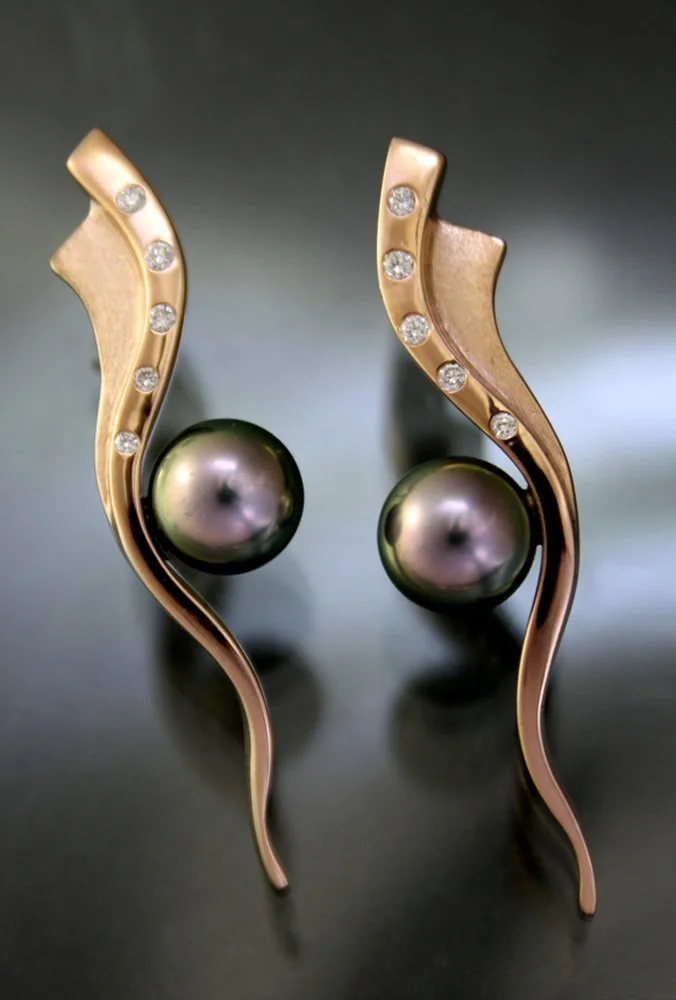 A pair of gold earrings with a wavy design, each featuring a large dark pearl and small diamonds along the curve.