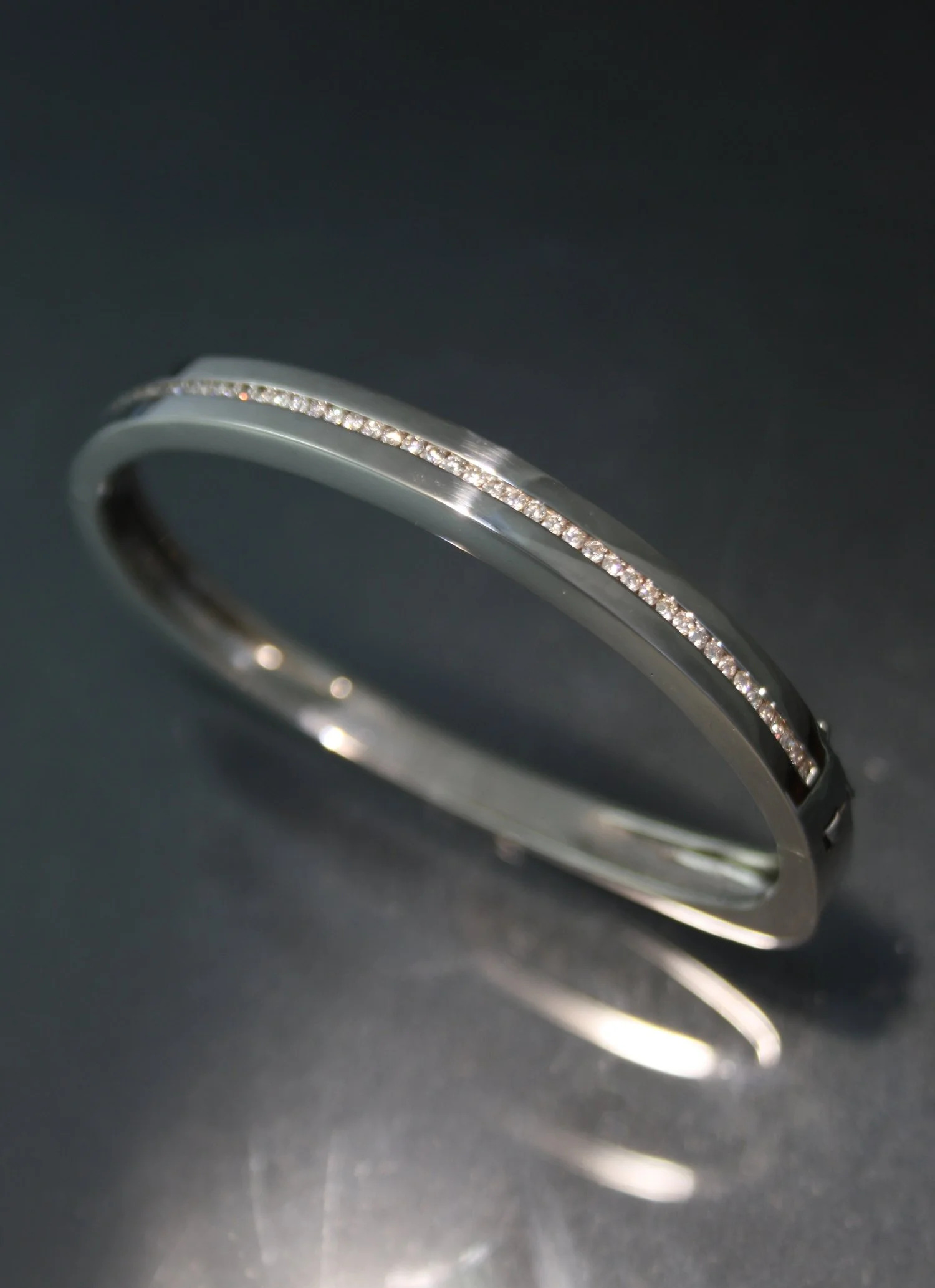 A silver bracelet with a line of small diamonds set along its top edge, resting on a dark reflective surface.