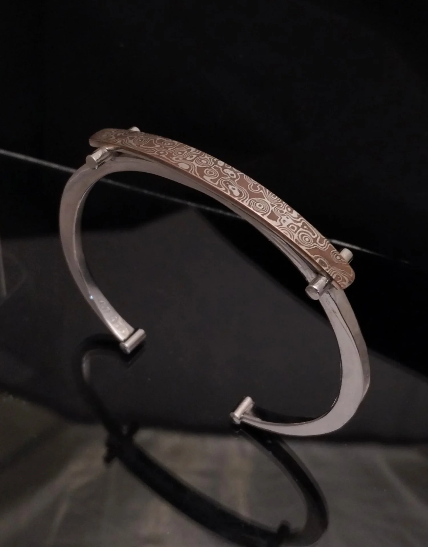 A stylish bangle bracelet with a copper-colored top featuring intricate swirl and geometric patterns, resting on a black reflective surface.