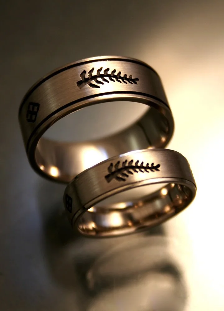 Two metallic wedding bands with engraved baseball and baseball stitching pattern, placed on a reflective surface with subtle lighting.