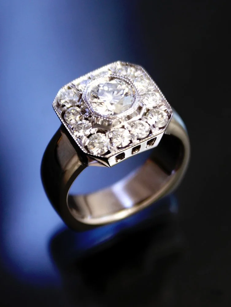 A silver ring with a large central diamond surrounded by smaller diamonds on a dark reflective surface.