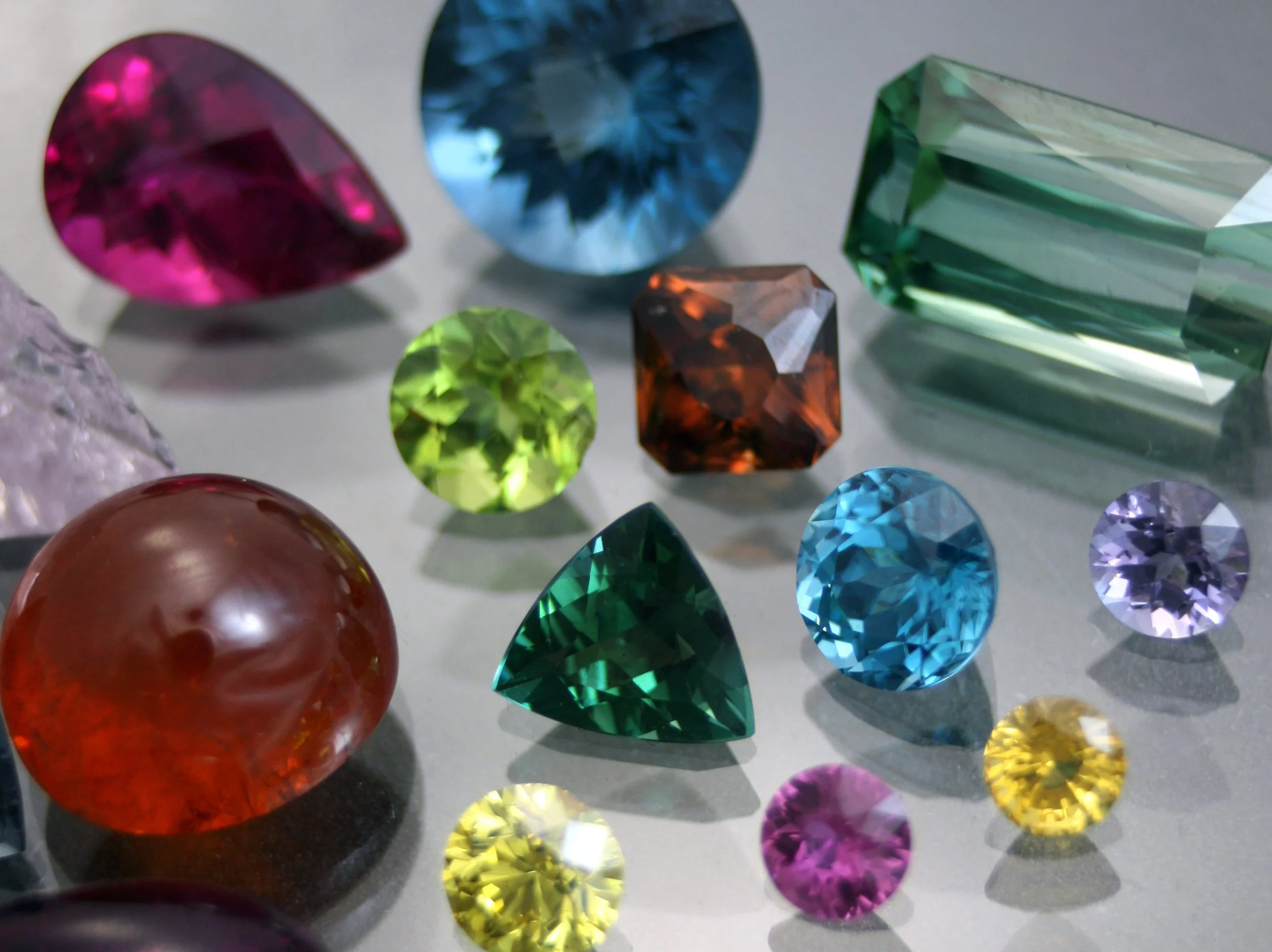 A collection of various colorful gemstones, including pink, blue, green, orange, purple, and yellow, in different shapes and sizes arranged on a gray surface.