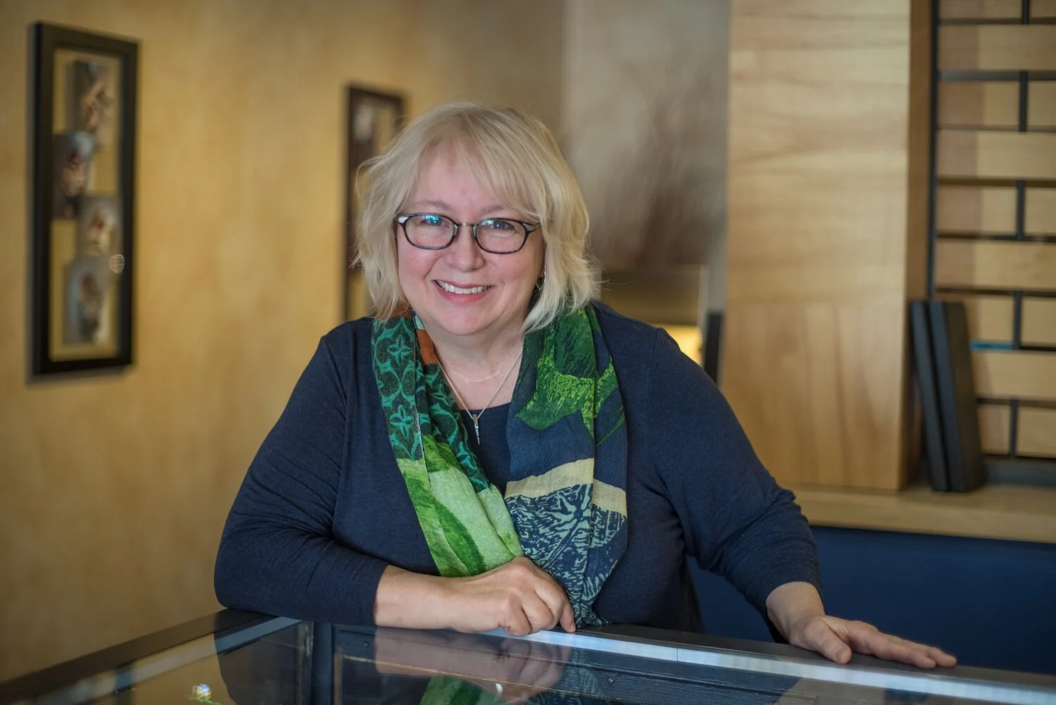 A woman with blonde hair, glasses, and a colorful scarf sitting at a table, smiling at the camera in a warmly lit room with framed pictures on the wall behind her.