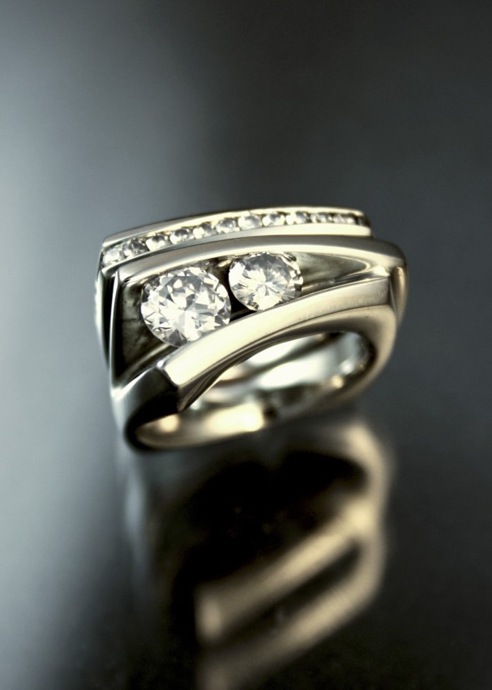 Silver ring with two large diamonds and smaller diamonds along the band, reflected on a dark surface.