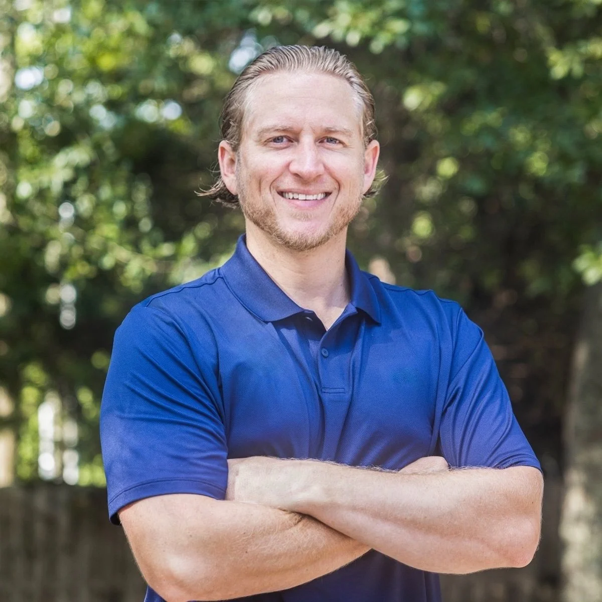 A smiling man with blonde hair and a beard, wearing a blue Under Armour polo shirt, standing outdoors with arms crossed in front of a tree-lined background.