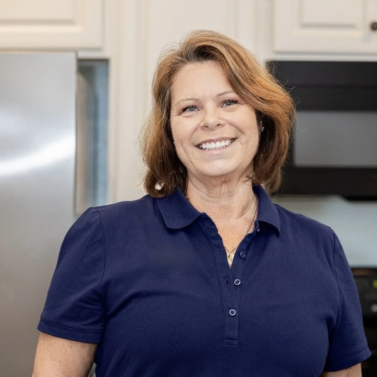 A woman with shoulder-length brown hair wearing a navy blue polo shirt with a logo on the chest, smiling in a kitchen with white cabinets and stainless steel appliances.