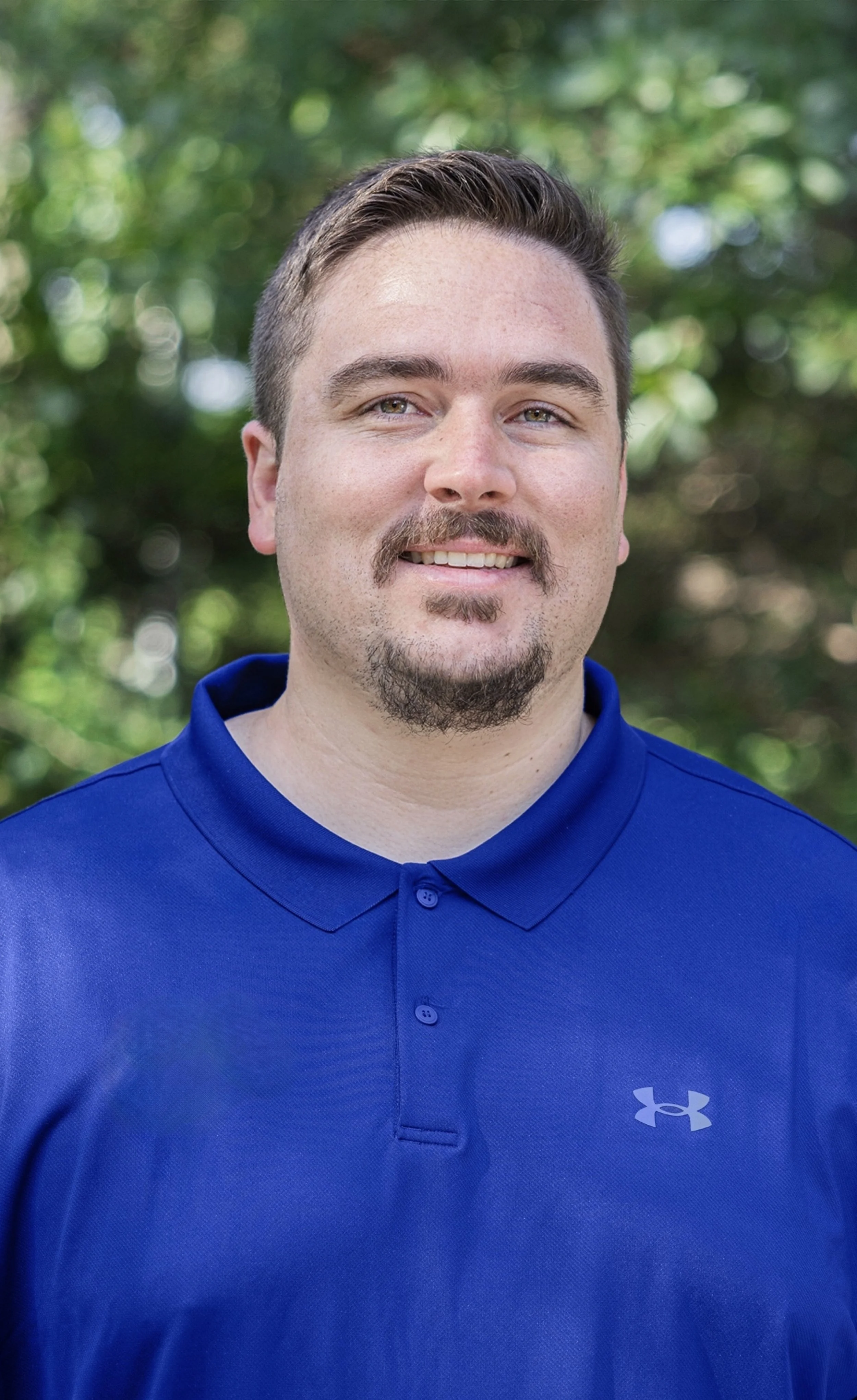 A man with short brown hair, a goatee, and a slight smile, wearing a navy polo shirt with a patch of a moon with a rising sun and a bow, standing in a kitchen with white cabinets and a black microwave.