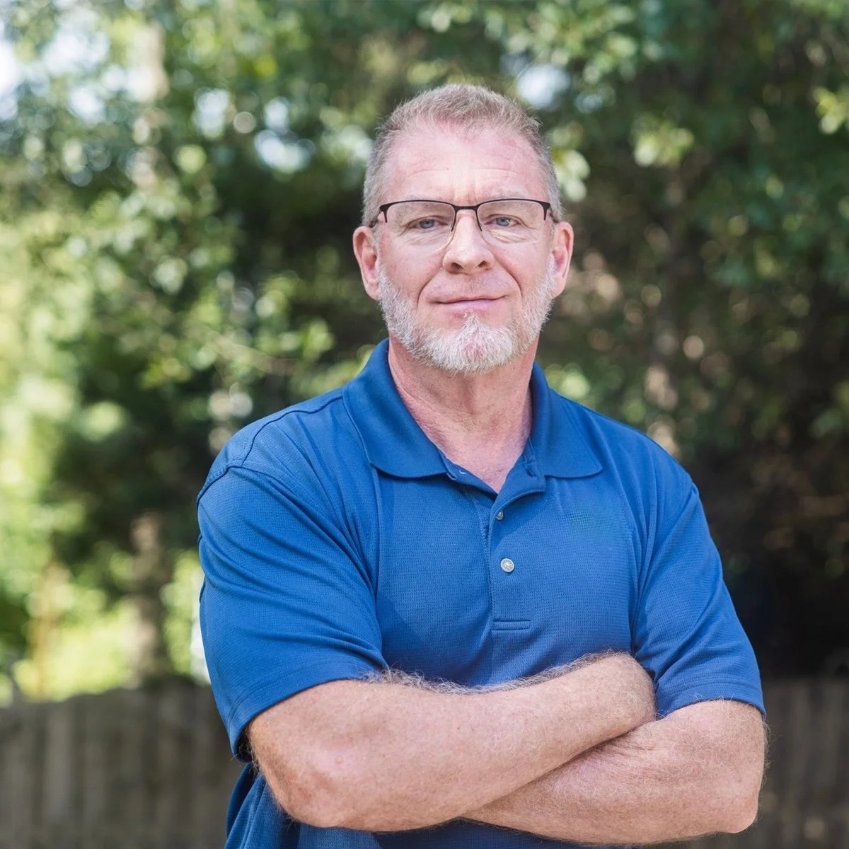 Portrait of a middle-aged man with glasses, a beard, and light-colored hair, standing outdoors with arms crossed, wearing a blue polo shirt with a logo, in front of a blurred background of trees.