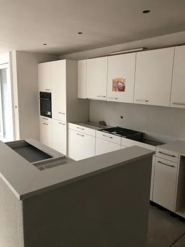 Moderne, weiße Küchenschränke in einer Einbauküche, helle Wände, integrierter Ofen und Herd, Küchenarbeitsplatte, kleine Fenster, Deckenleuchten.