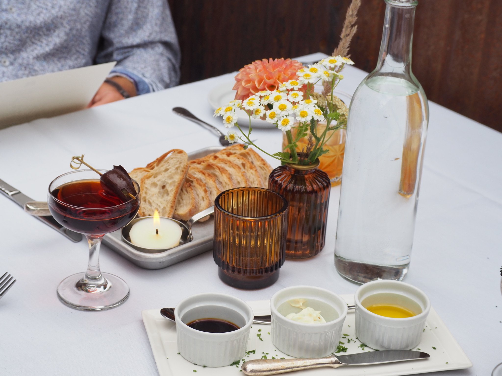 A table set for a meal with sliced bread, a glass of red wine with a cherry garnish, small bowls of condiments, a bottle of water, a small bouquet of daisies and other flowers in a brown vase, candles, and a person in a gray sweater sitting at the table.