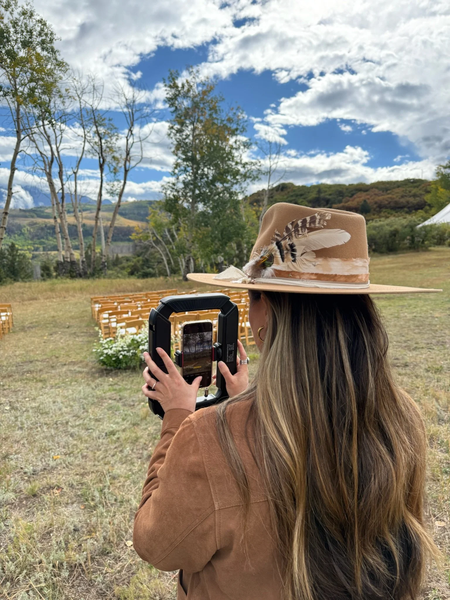 A woman with long, light brown hair wearing a tan hat adorned with feathers, taking a photo using a phone and a stabilizer, outdoors in a grassy area with chairs and trees in the background, under a partly cloudy sky.