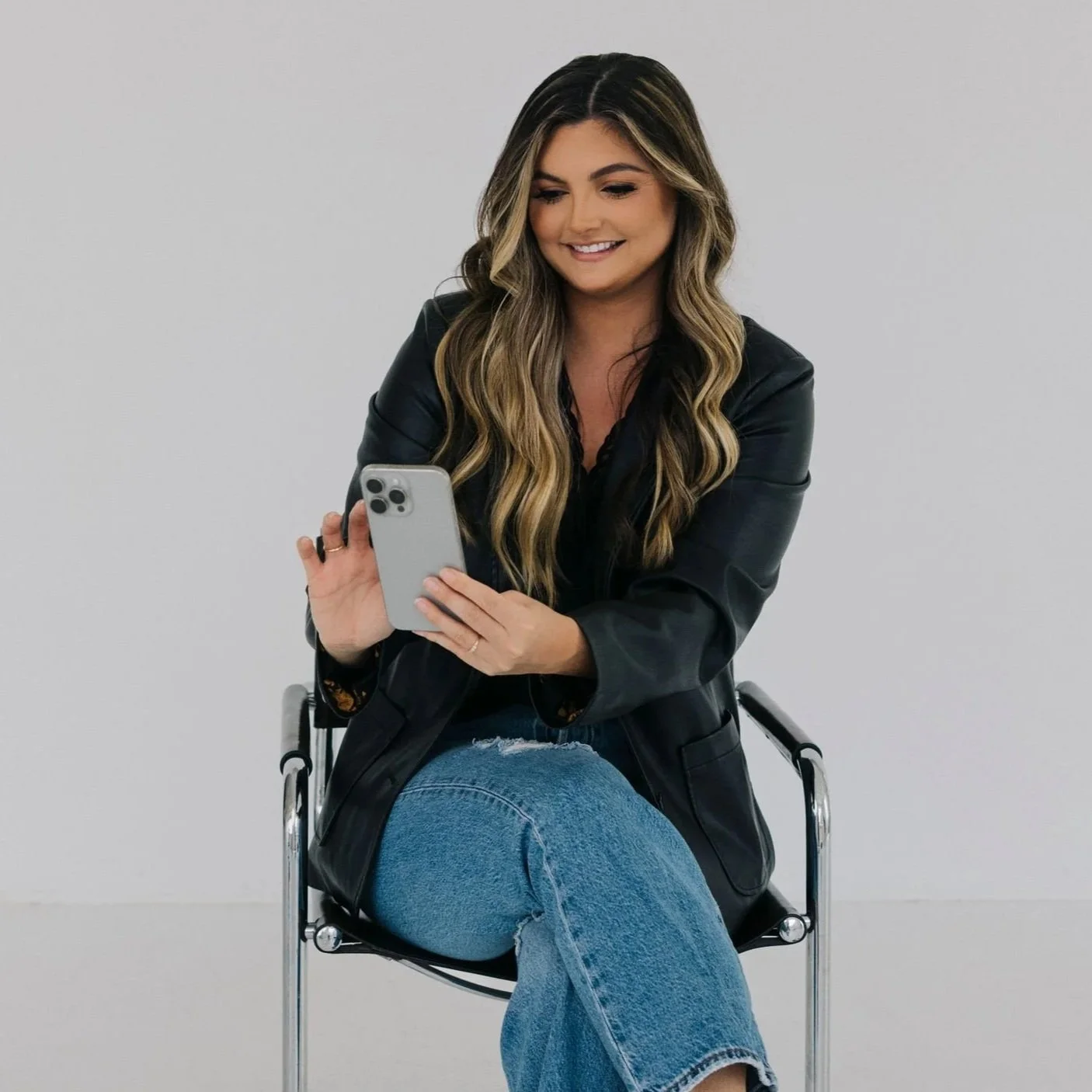 A young woman with long wavy hair, wearing a black leather jacket and blue jeans, sitting on a modern chair, looking at her smartphone with a smile.