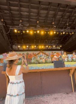 A woman wearing a hat and white dress with a belt stands in front of an outdoor stage, taking a photo of the stage with her phone. The stage has colorful artwork painted on its backdrop and is illuminated by yellow stage lights.