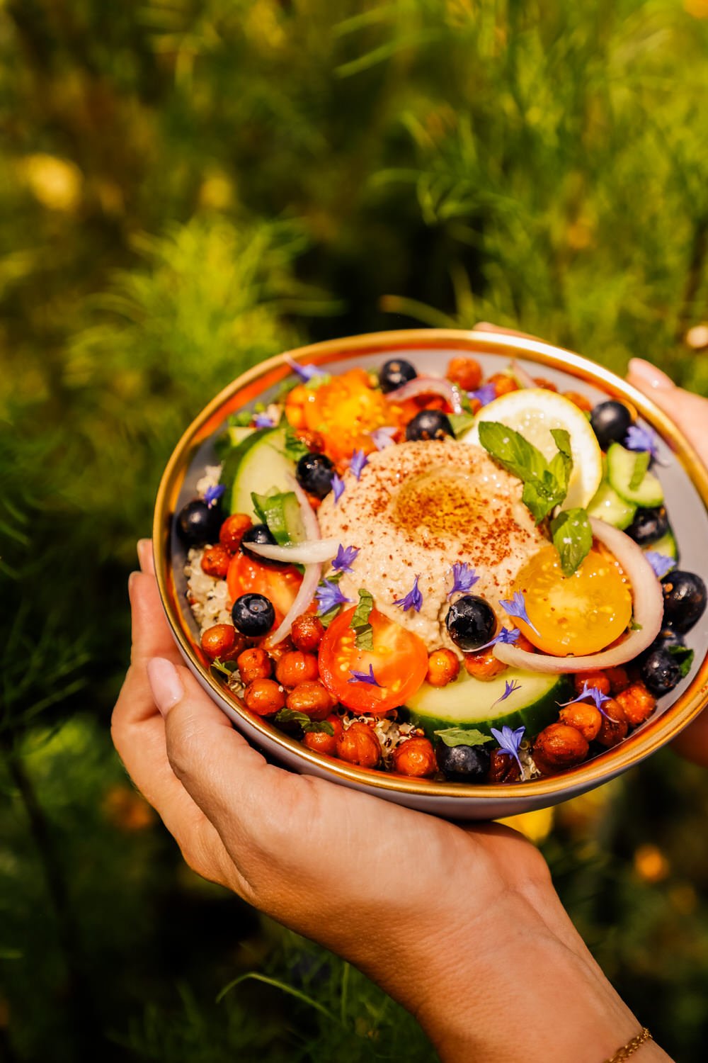 A bowl of Greek salad with cucumbers, tomatoes, red onion, chickpeas, blueberries, and herbs, topped with hummus and sprinkled with paprika.