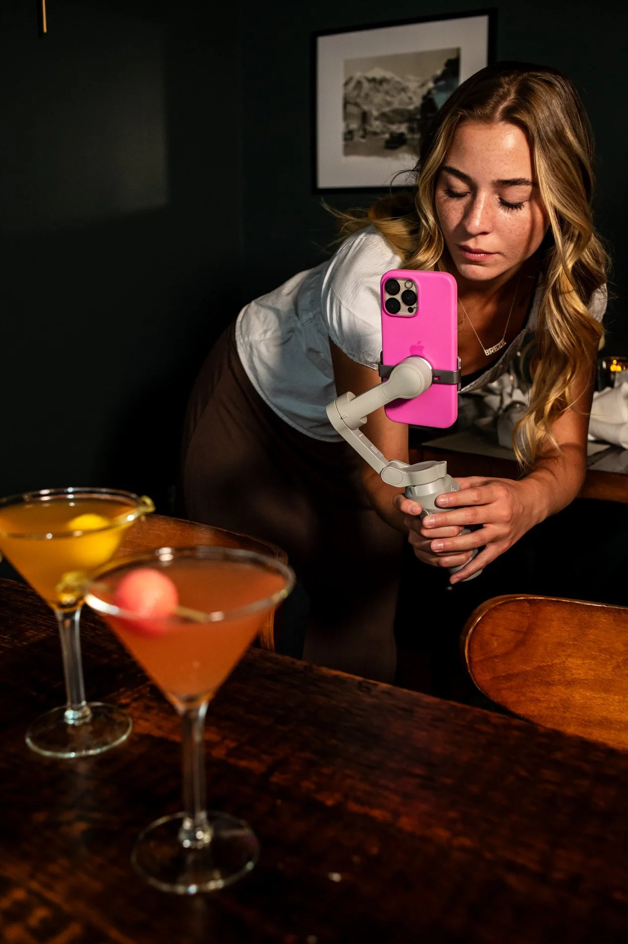 Woman taking a photo with a pink smartphone mounted on a stabilizer, leaning over a table with two cocktails topped with fruit garnishes.