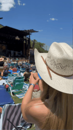 Person wearing a wide-brimmed hat, sitting outdoors at a music festival or concert during daytime, surrounded by an audience on chairs, with a stage in the background and a clear blue sky.