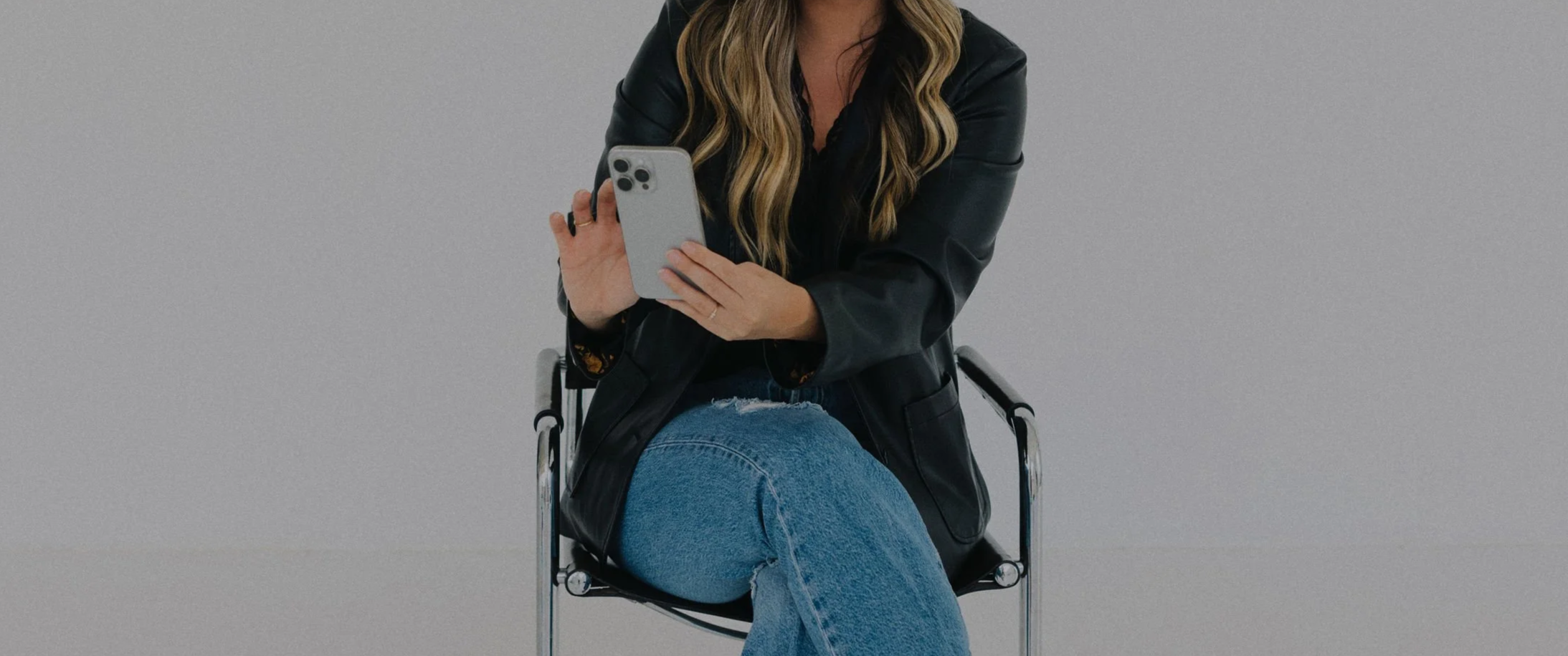 A woman with long wavy hair, wearing a black leather jacket and ripped blue jeans, sitting on a black chair, looking at her smartphone held in her left hand against a plain gray background.
