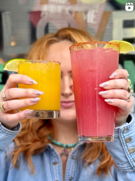 A woman with red hair holding a yellow smoothie and a pink smoothie, each garnished with a lime wedge, in a casual outdoor setting.