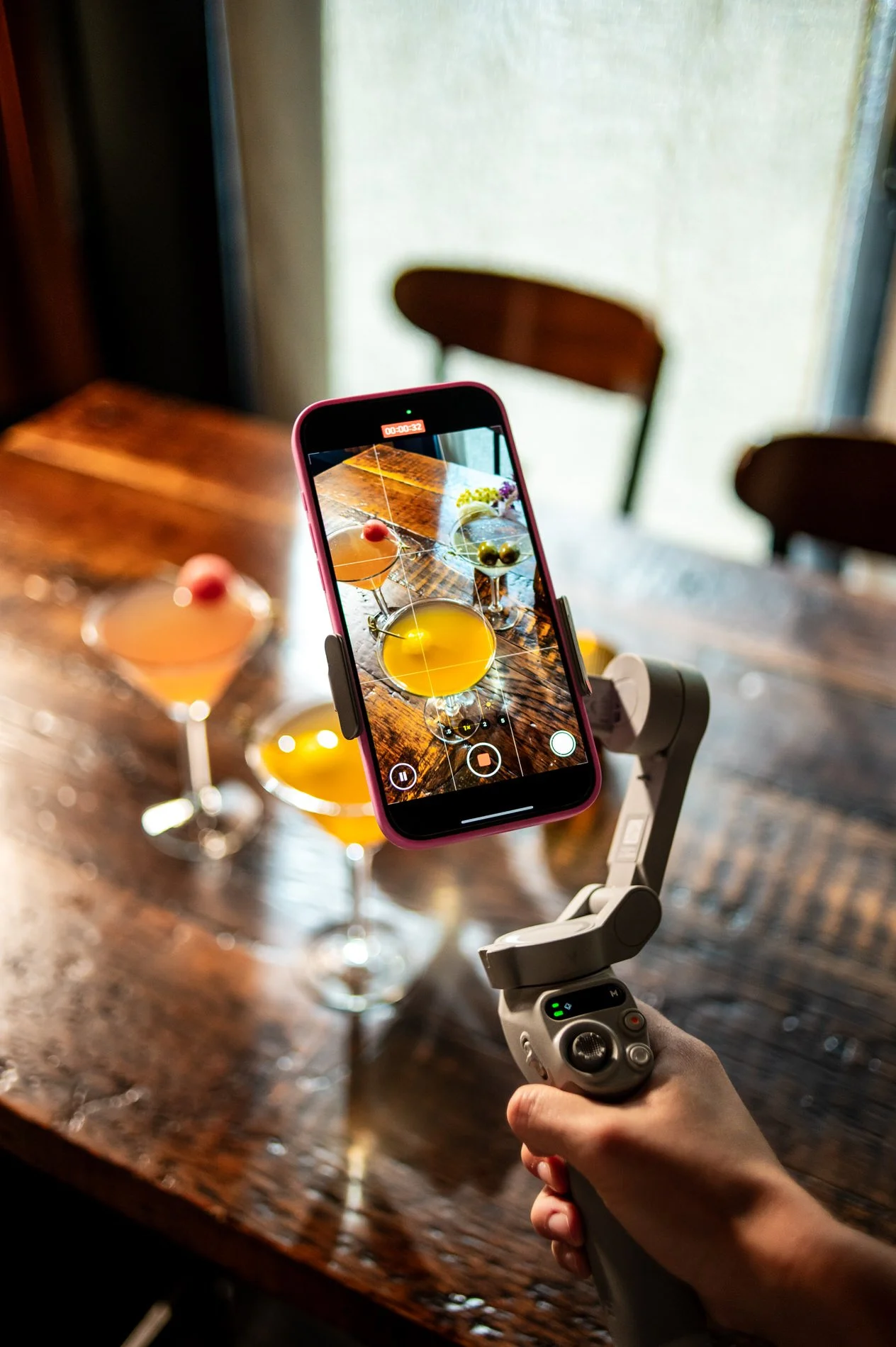 A person holding a camera stabilizer capturing photos of four cocktails with colorful garnishes on a wooden table near a window.