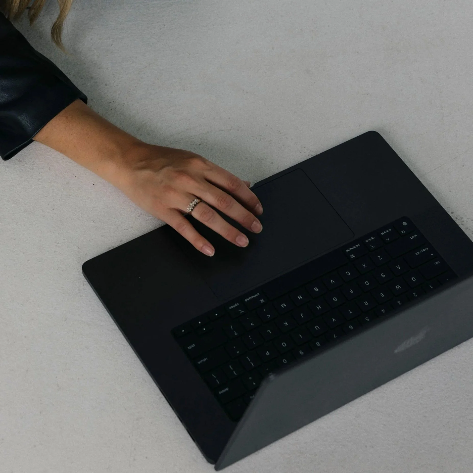 A person's hand with a ring on the ring finger resting on a black laptop keyboard on a light-colored surface.