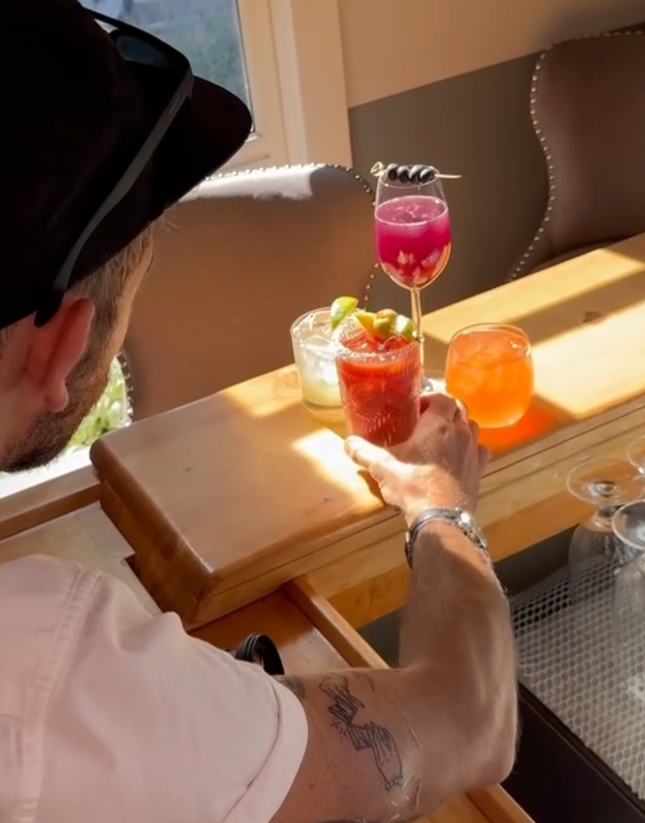 A man wearing a cap and bracelet preparing various colorful cocktails on a wooden serving board in a well-lit room with chairs.