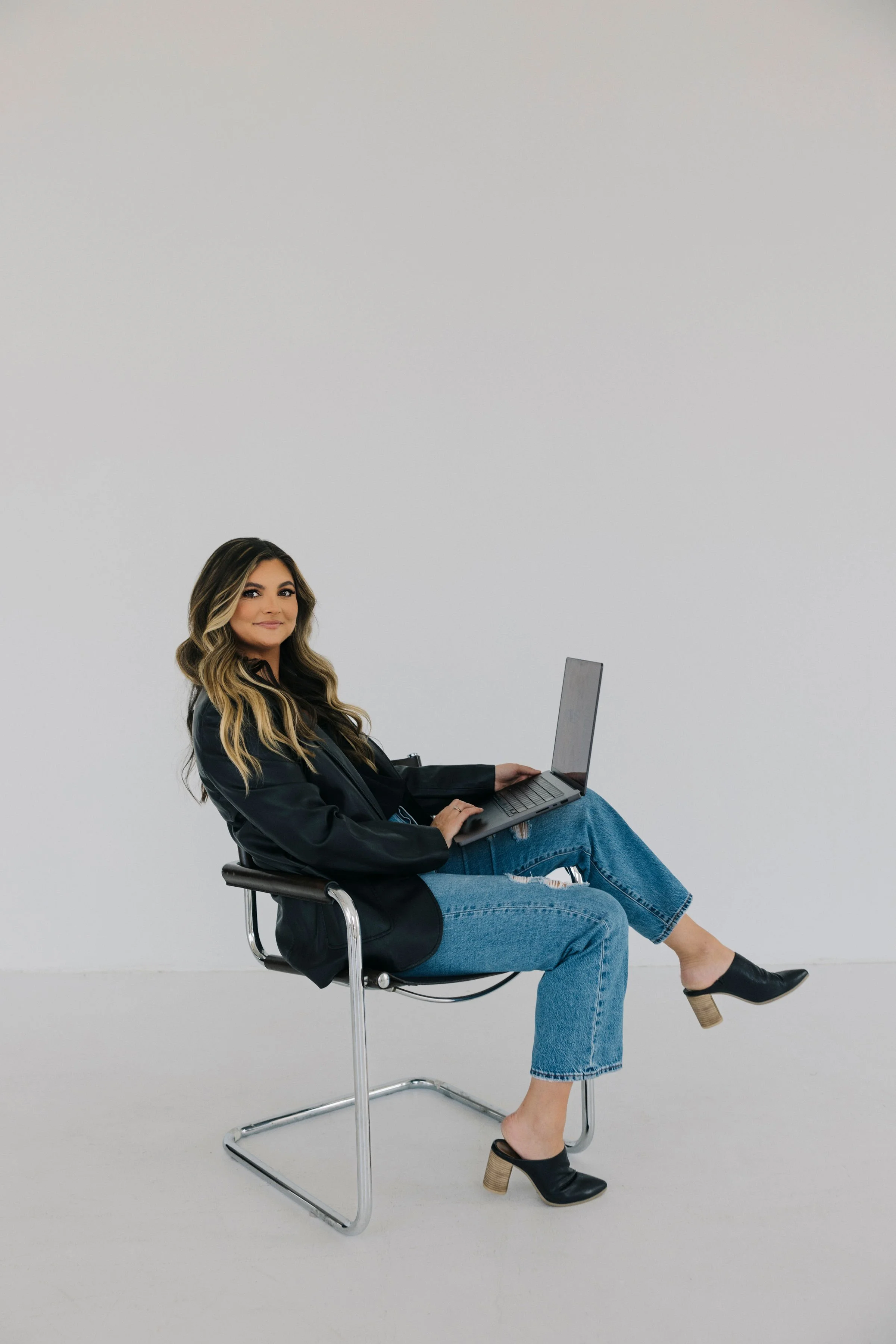 Woman sitting on a modern chair, working on a laptop in an empty white room.