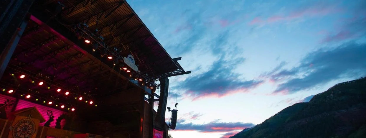 An outdoor stage at a festival at dusk with colorful lights and mountains in the background.