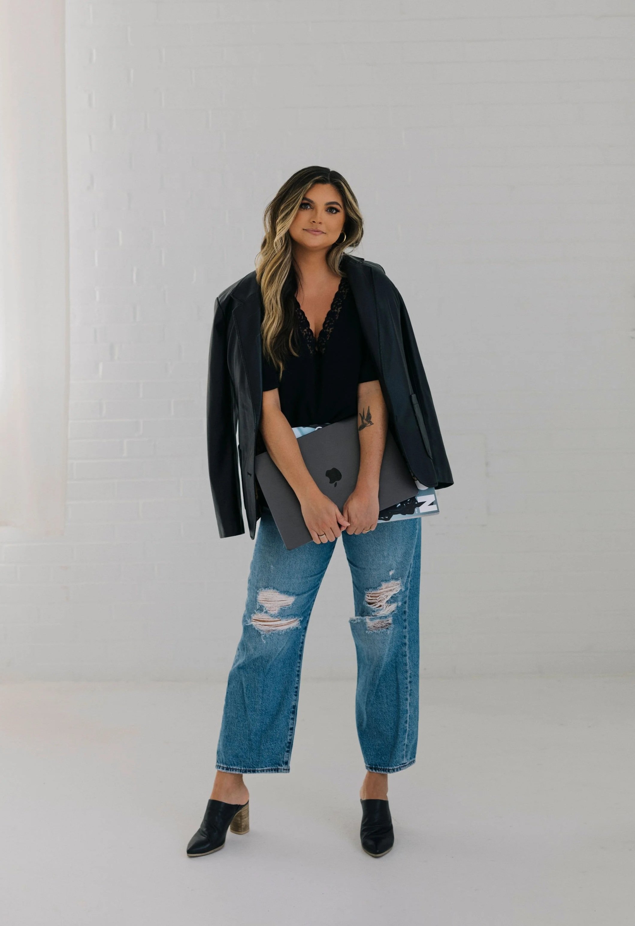 Woman standing against a white brick wall, holding a closed laptop, dressed in a black top, distressed jeans, black boots, and a black jacket draped over her shoulders, with wavy hair and makeup.