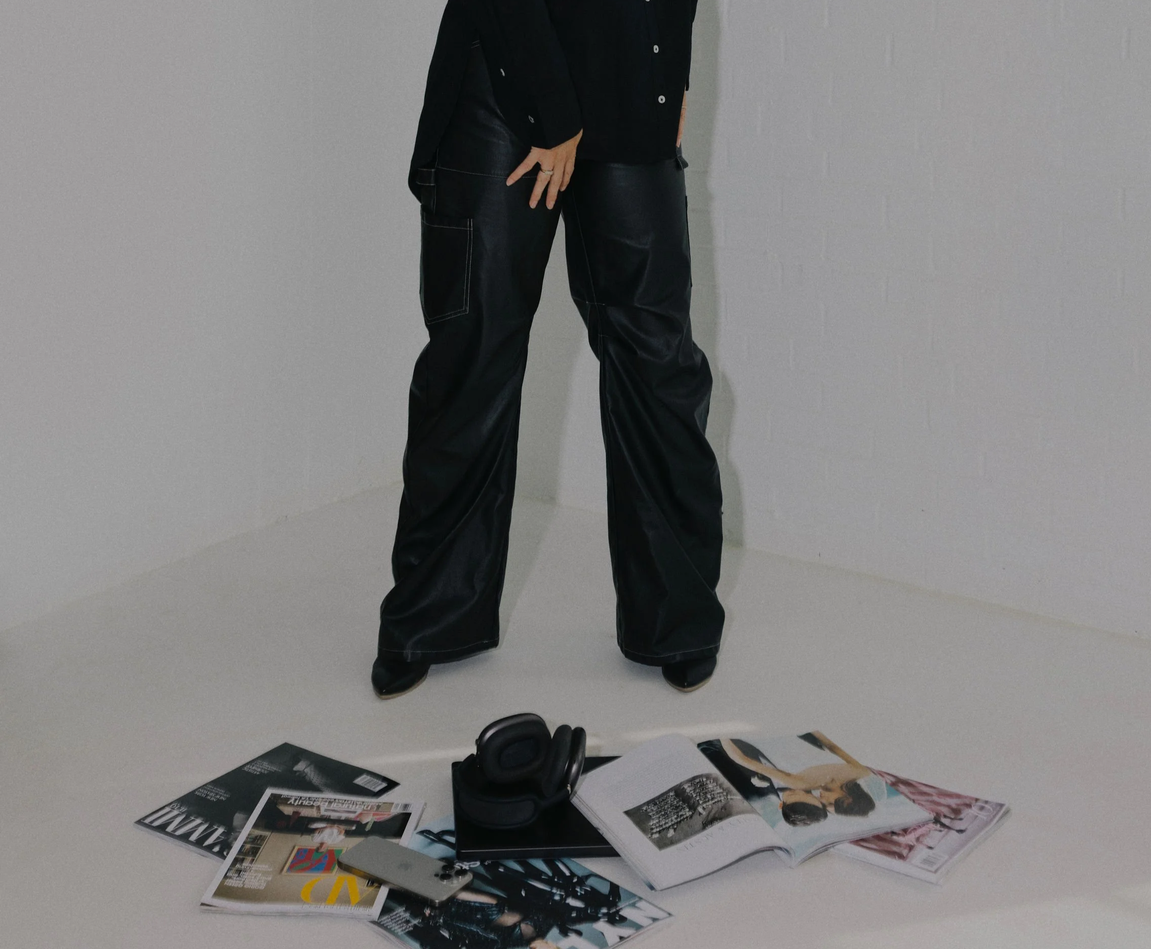 Partially visible person standing in a white room, wearing black pants and black shoes, with magazines, a pair of headphones, and a remote on the floor.