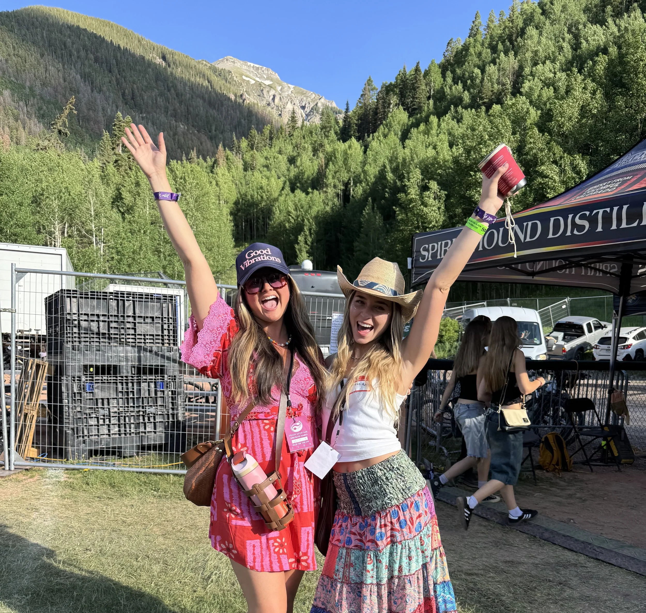 Two women celebrating outdoors at a festival or event in front of a forested mountain landscape, smiling with arms raised, holding a red tumbler, wearing hats and festival wristbands.
