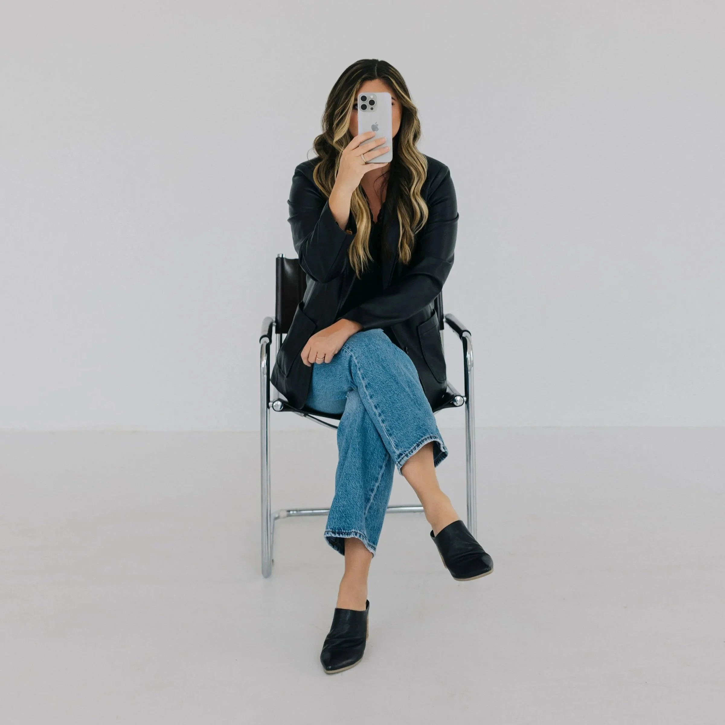 Woman with long wavy hair taking a selfie with an iPhone, sitting on a black chair, wearing a black blazer, blue jeans, and black heels in a minimalist space.