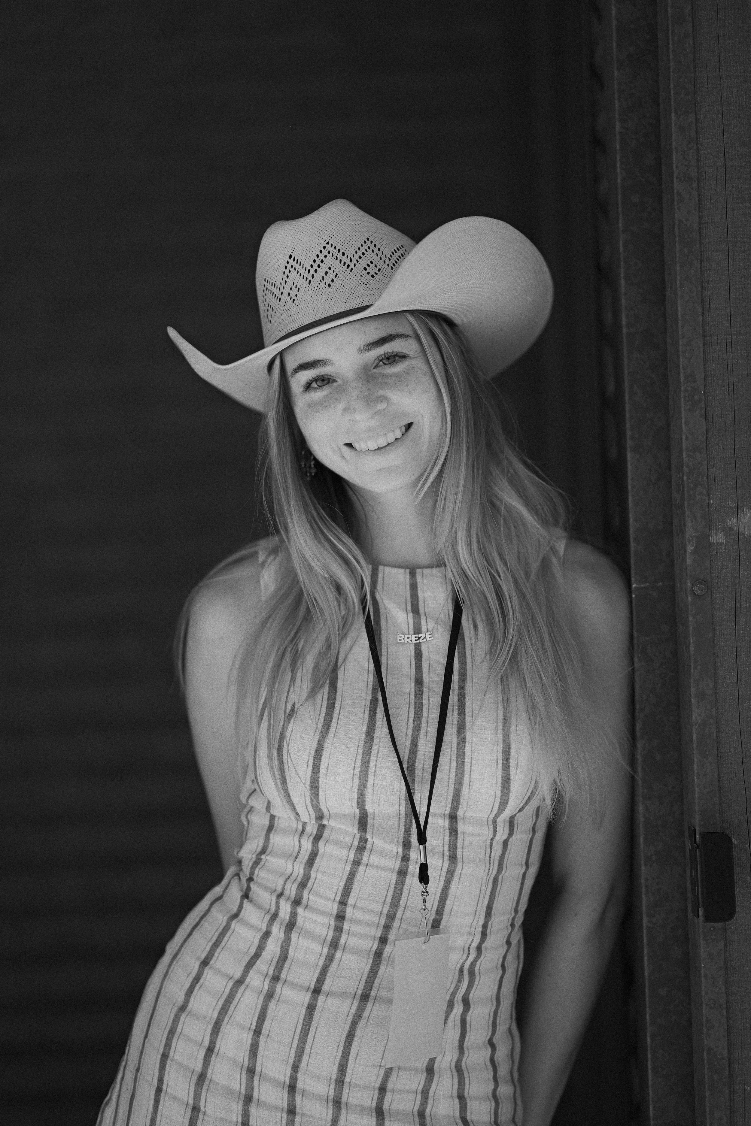 A young woman with long hair smiling, wearing a wide-brimmed cowboy hat, a striped sleeveless dress, and a necklace with the name "BREEZE" on it, standing near a wooden door.