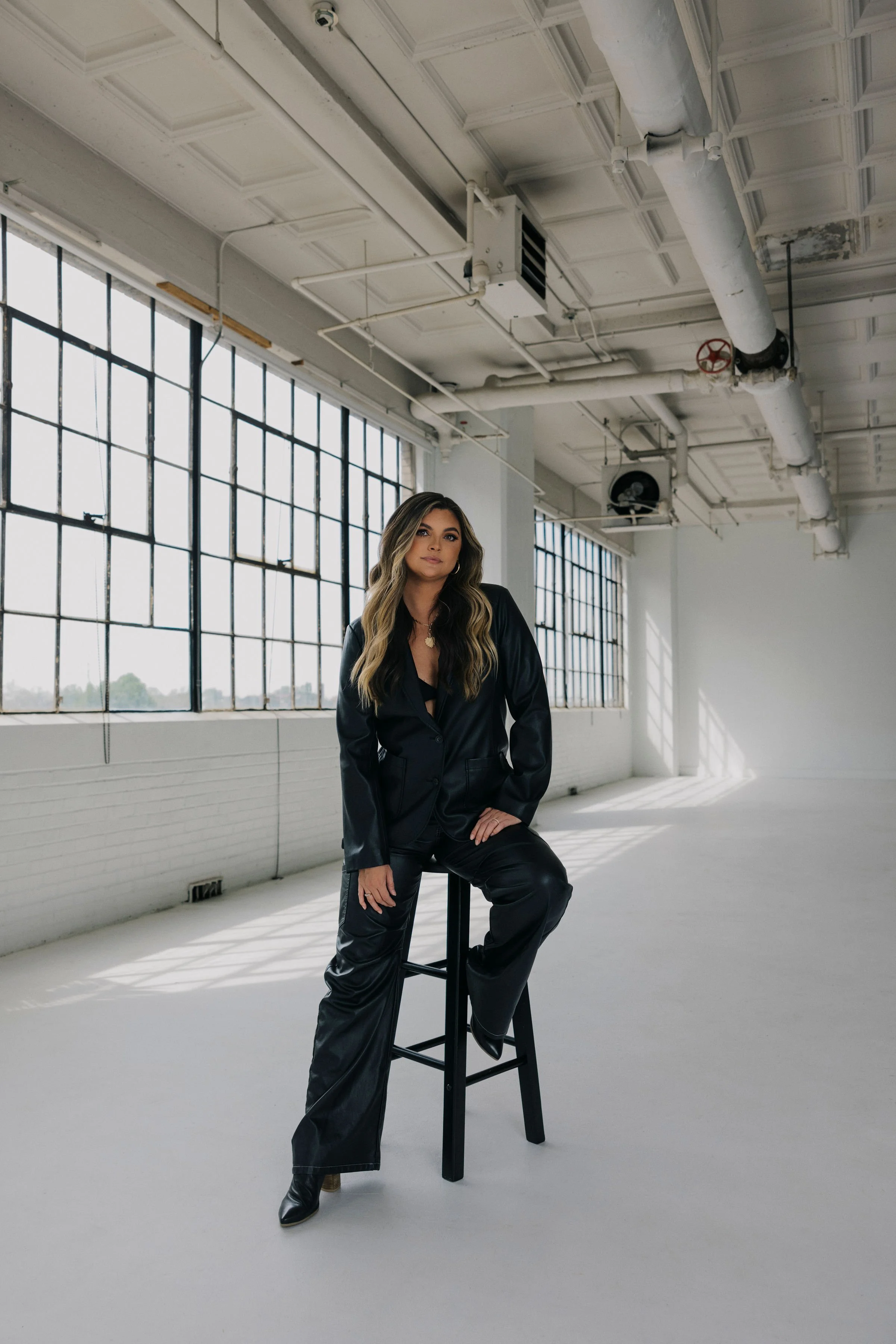 A woman with long wavy hair wearing a black leather suit and black boots sitting on a tall black stool in a spacious industrial-style studio with large windows, white walls, and exposed ceiling pipes.