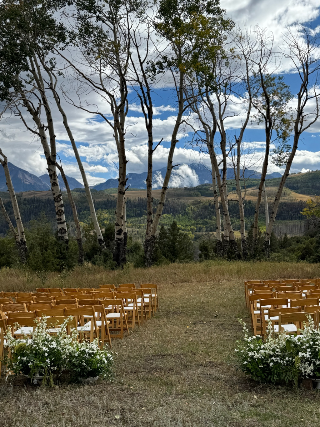 Outdoor wedding ceremony setup with wooden chairs arranged on grass, white floral arrangements in the foreground, tall leafless trees, green hills, mountains, and partly cloudy sky in the background.
