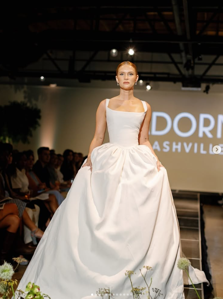 A model walking on a runway during a fashion show, wearing a white wedding gown with a fitted bodice and full skirt, with audience members seated on either side and a large illuminated sign in the background reading 'NORDSTROM NASHVILLE.'