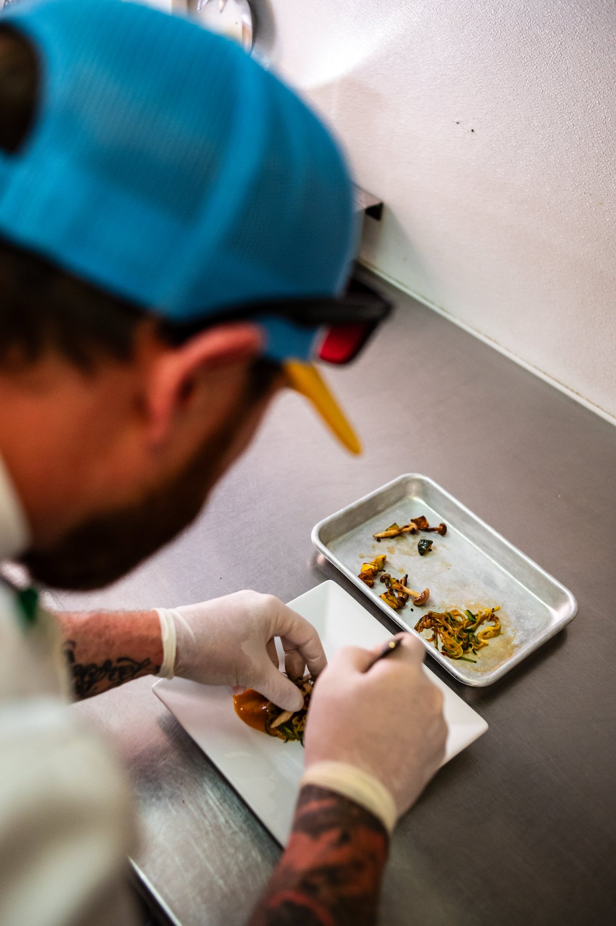 A chef wearing gloves and a blue cap is carefully plating cooked vegetables onto a white dish in a professional kitchen.