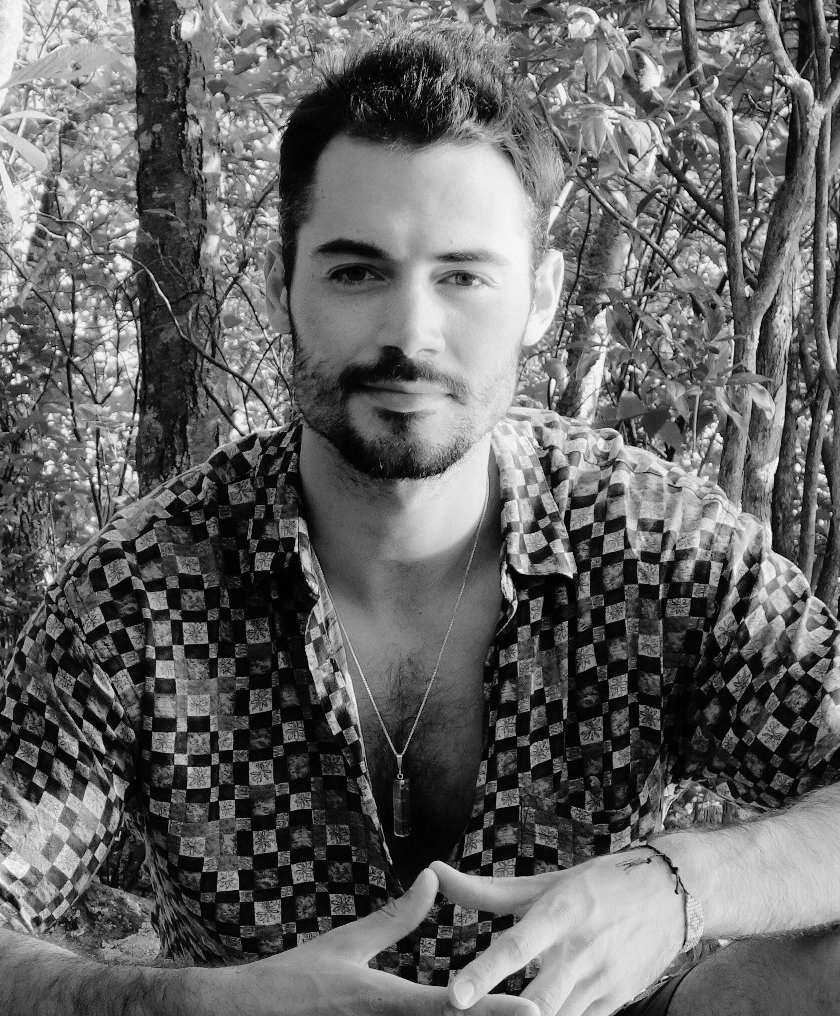 A young man with short dark hair, a beard, and mustache, wearing a checkered shirt with the top buttons undone, a necklace with a pendant, and a bracelet, sitting outdoors with trees and foliage in the background.