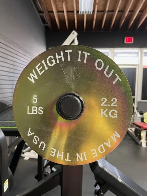 A weight plate with a humorous message that reads "Weight it out, made in the USA," weighs 5 pounds (2.2 kg), and is located inside a gym.