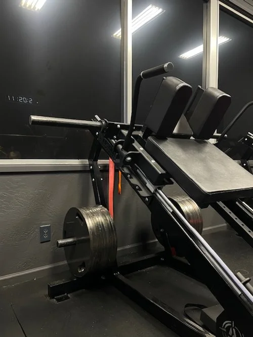 A weight bench with black padding, angled incline, multiple seated positions, in front of a large window with a black wall and floor, and three seated machines visible in the background.