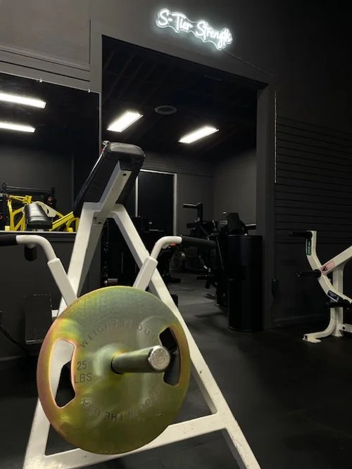 Indoor gym with workout equipment including a barbell on a squat rack, weighing 25 pounds, in the foreground. There are various machines and weights in the background under bright ceiling lights.