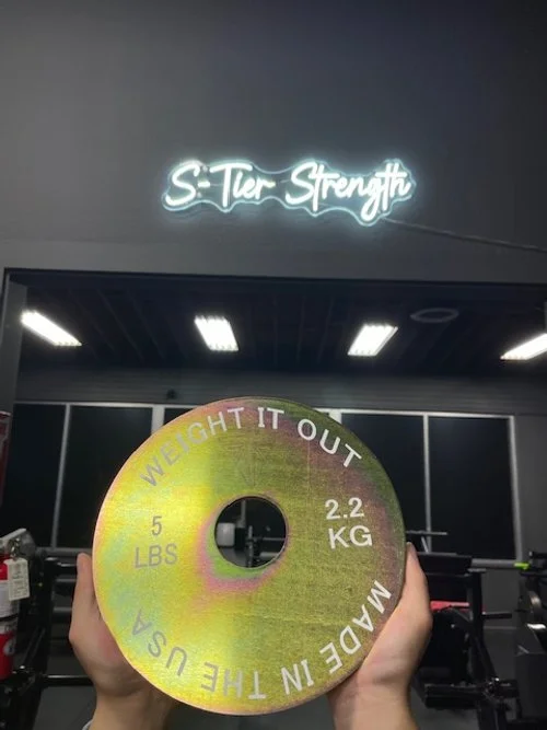 A person holding a weight plate with the text 'Weight It Out' and 'Made in the USA' printed on it, with a neon sign above reading 'S-Ter Strength' in a gym.