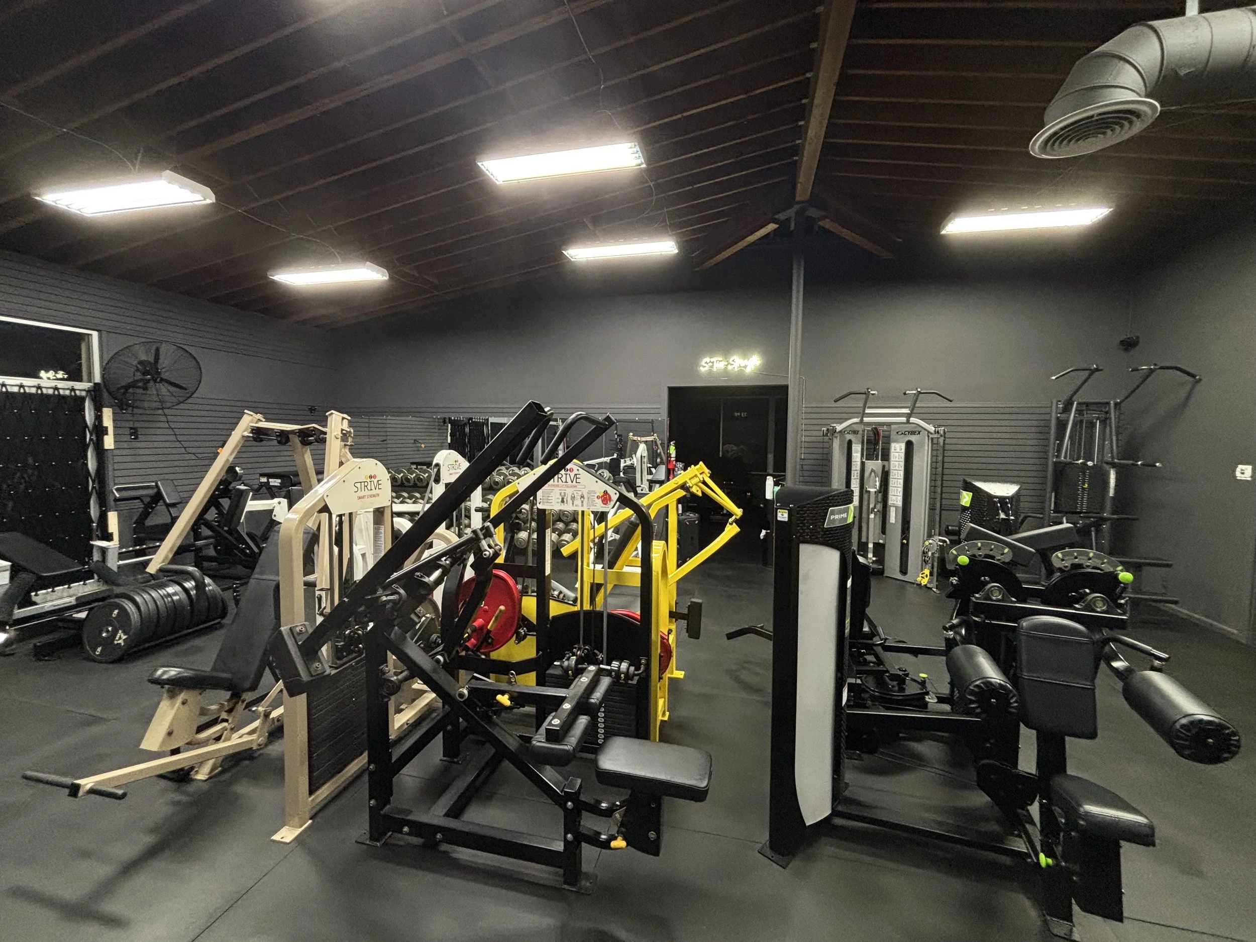 Empty gym with various workout equipment, including weight machines, free weights, and cardio machines, in a modern fitness center with dark walls and ceiling.