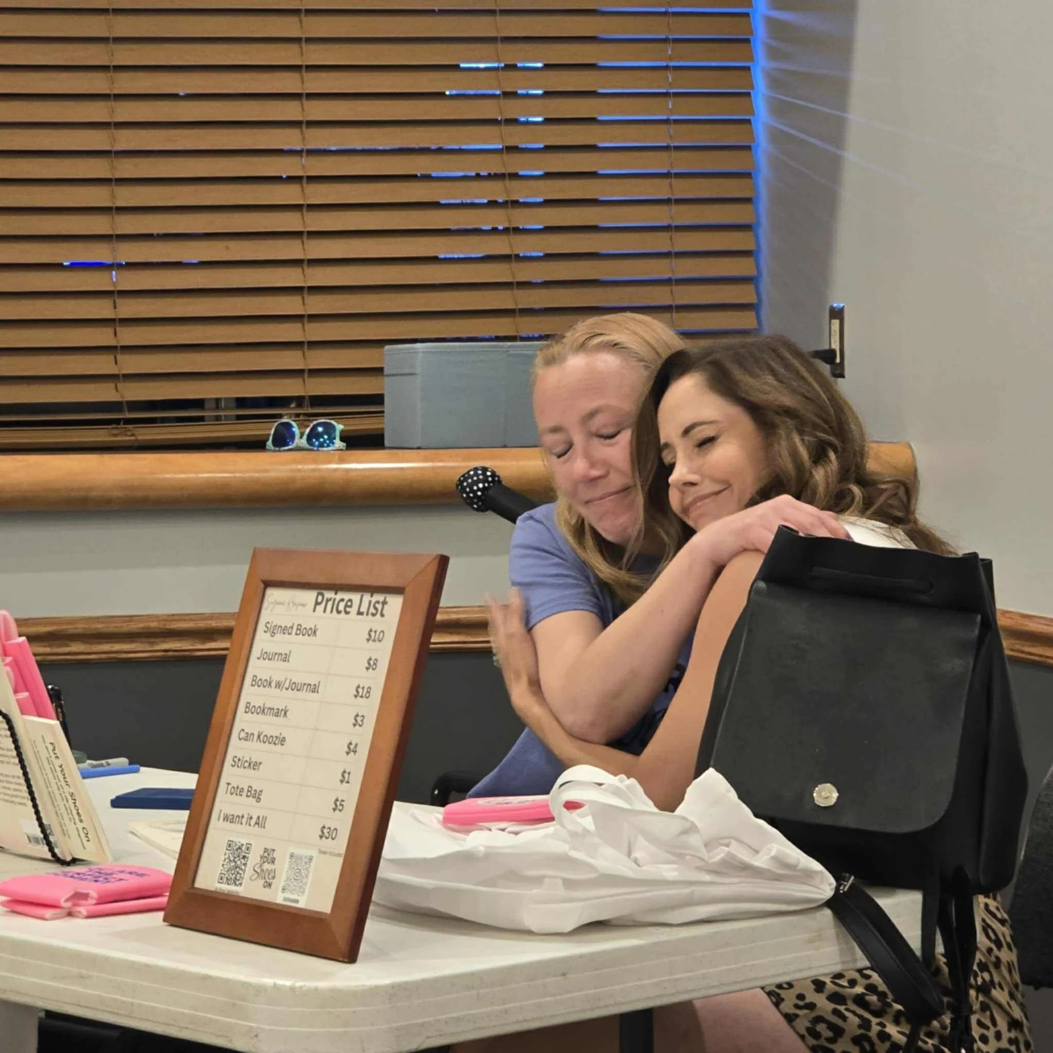 Two women embrace at a table, smiling with eyes closed. The table has pink pens, a black purse, and a framed price list with various items and prices, including a signed book for $10 and a tote bag for $5.