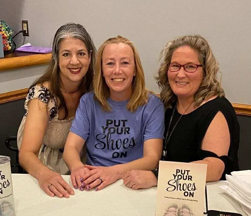 Three women sitting at a table, smiling, with a sign that reads 'Put Your Shoes On' on the table.