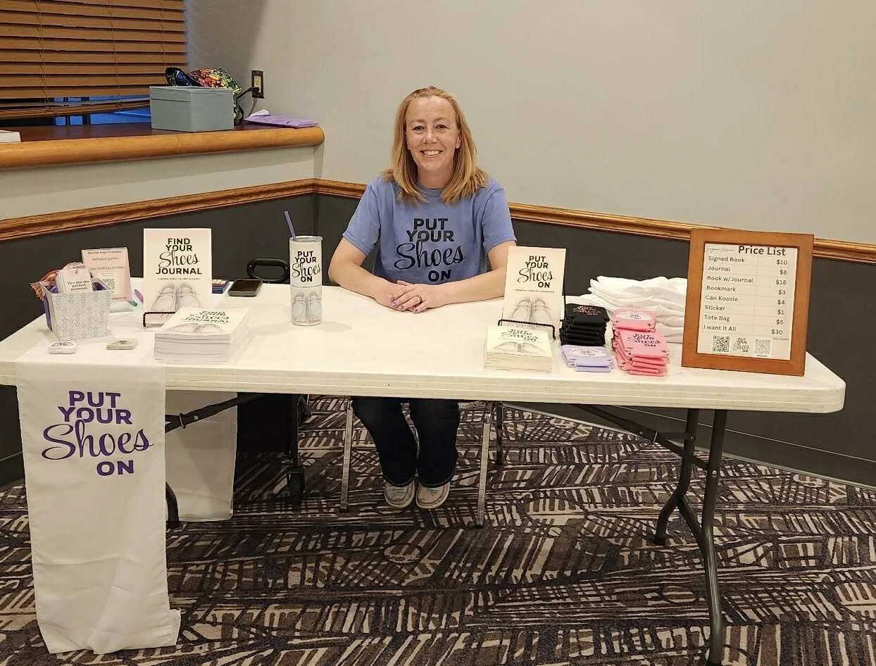 Woman sitting behind a table at a shoe-related sales booth with a sign that says "PUT YOUR Shoes ON" and various items for sale including books, stickers, and tote bags, with a price list on a framed board.
