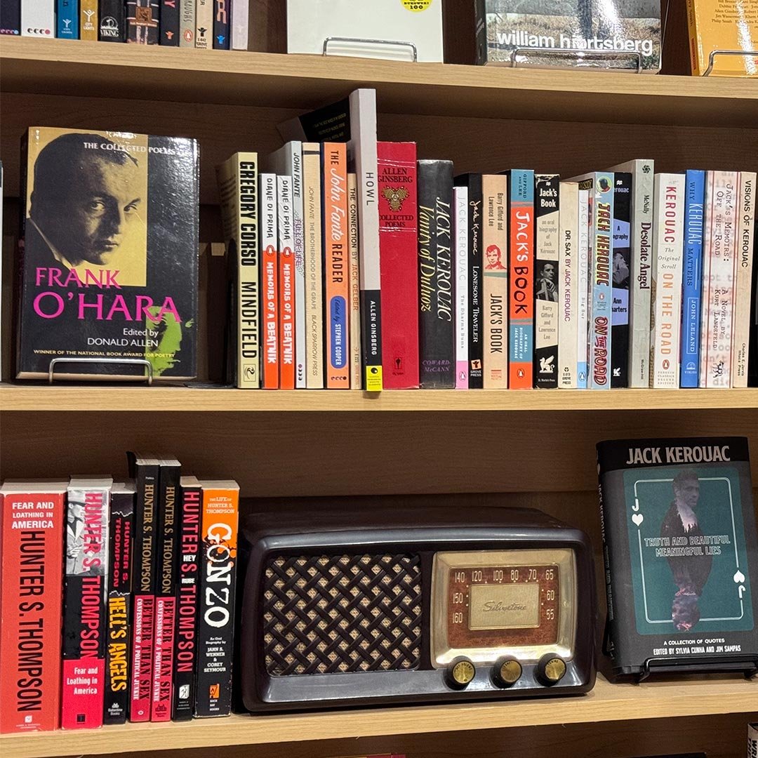 Bookshelf with various books, including works by Frank O'Hara, J.K. Rowling, Jack Kerouac, and others. An antique radio is on the shelf below the books.