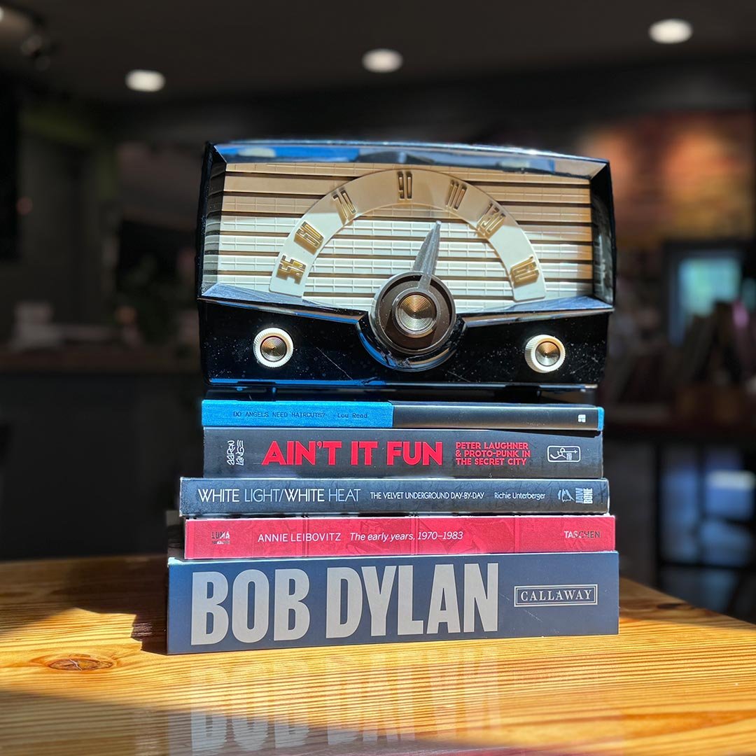 Stack of five books on a wooden table with a vintage radio on top. The books are titled "Ain't It Fun," "White Light/White Heat," "Annie Leibovitz," and "Bob Dylan." The background shows a blurred indoor setting with warm lighting.