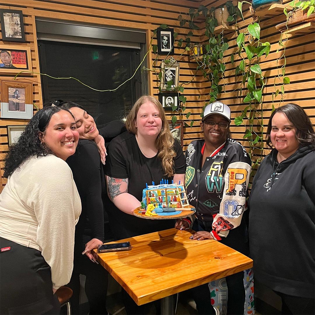Five women celebrating around a birthday cake with a library theme, indoors with wooden walls, framed photos, and green plants.
